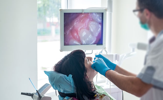 Dentist examining patient's mouth with endoscope while displaying internal imagery on monitor above.