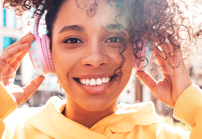 Young girl smiling brightly wearing yellow hoodie with hands near face in sunny outdoor setting.