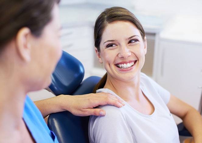 Female dentist in white coat smiling at patient in dental office chair during consultation.