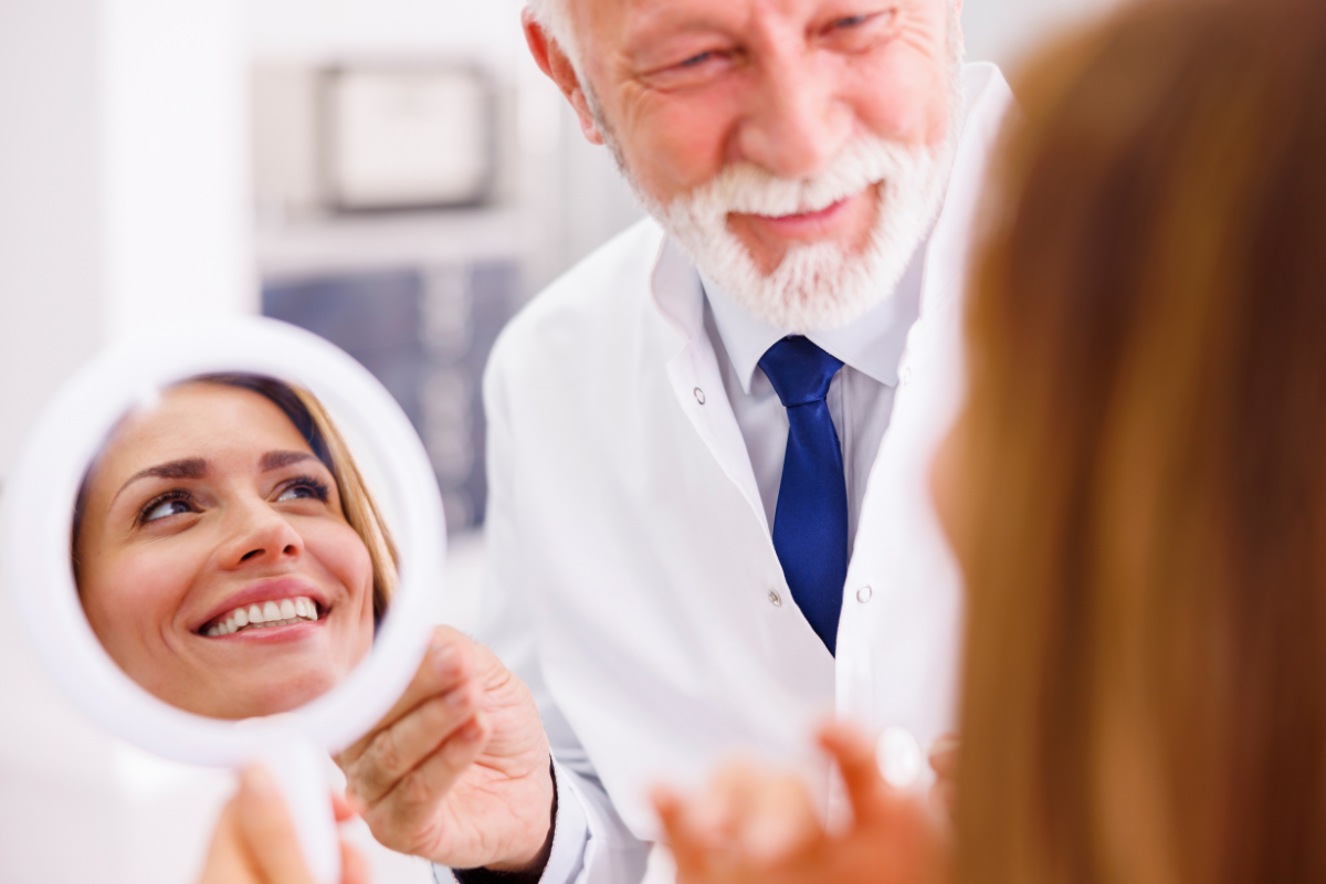 Older male dentist showing female patient her smile in handheld mirror in dental office.